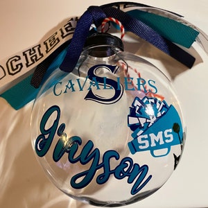May include: A clear glass ornament with a teal and white design. The ornament has the text "CAVALIERS" and "SMS" on it. The name "Grayson" is written in teal cursive on the ornament.