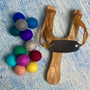 May include: A wooden slingshot with a brown leather pouch and a set of colorful felt balls. The felt balls are in various shades of blue, green, red, purple, pink, and yellow.