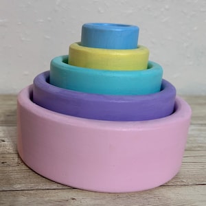 May include: A pastel-colored wooden stacking toy with a pink base, followed by purple, teal, yellow, and blue rings. The toy is designed to nest inside each other, creating a tower. The toy is on a light wood surface.