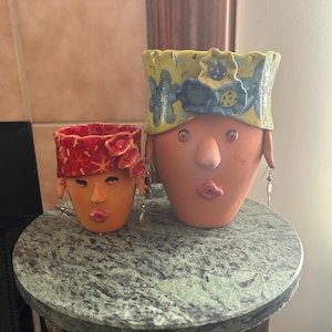 May include: Two terracotta face planters with floral headbands and dangling earrings. The larger planter has a yellow headband with blue and white flowers. The smaller planter has a red headband with a floral design. Both have painted facial features.