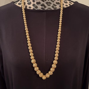 May include: A long gold-colored necklace with intricate, openwork spherical beads. The necklace is displayed against a black background, creating a contrast that highlights the jewelry's details. The beads vary in size, adding visual interest to the piece.