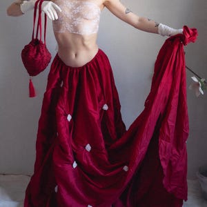 May include: A woman wearing a white lace bandeau top and a long, flowing burgundy skirt with white embellishments. She is holding a small, red, beaded bag with a tassel.
