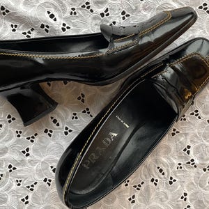 May include: A pair of black leather heeled loafers with gold stitching detail. The loafers have a pointed toe and a low block heel. The brand name "PRADA" is visible on the sole of one shoe.