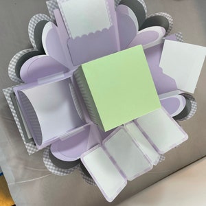 Pode incluir: Uma caixa de explos&atilde;o de cor lavanda e cinza com m&uacute;ltiplos compartimentos para fotos ou mensagens. A caixa tem um quadrado verde claro no centro.