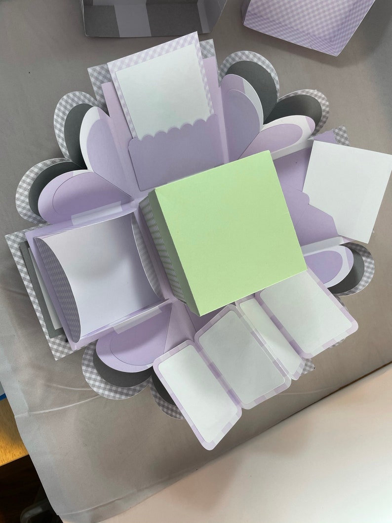 Pode incluir: Uma caixa de explos&atilde;o de cor lavanda e cinza com m&uacute;ltiplos compartimentos para fotos ou mensagens. A caixa tem um quadrado verde claro no centro.