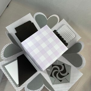 Pode incluir: Uma caixa de explos&atilde;o xadrez cinza e branca com uma caixa xadrez roxa e branca dentro. A caixa tem v&aacute;rios compartimentos com inser&ccedil;&otilde;es de papel preto e um design de redemoinho de papel preto.