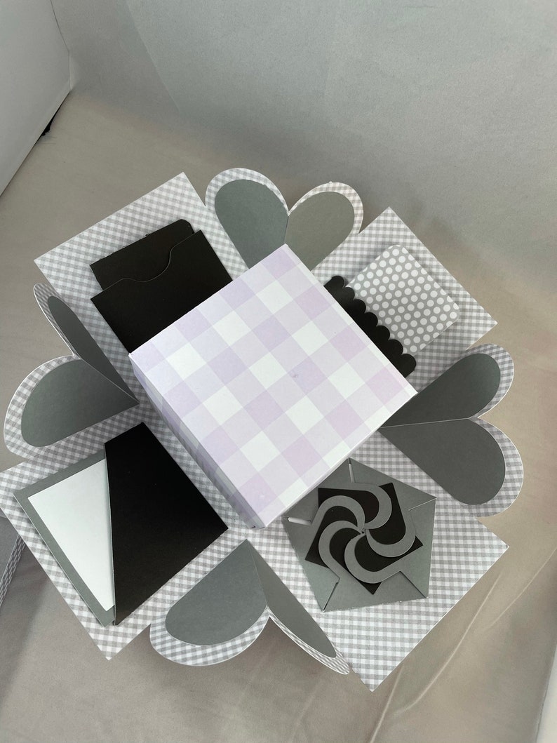 Pode incluir: Uma caixa de explos&atilde;o xadrez cinza e branca com uma caixa xadrez roxa e branca dentro. A caixa tem v&aacute;rios compartimentos com inser&ccedil;&otilde;es de papel preto e um design de redemoinho de papel preto.