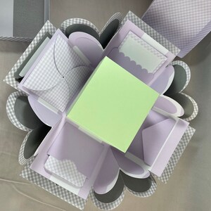 Pode incluir: Uma caixa de explos&atilde;o xadrez cinza e branca com um quadrado verde claro no centro. A caixa tem v&aacute;rias abas que se abrem para revelar diferentes compartimentos.