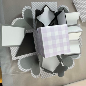 Pode incluir: Uma caixa de explos&atilde;o xadrez cinza e branca com uma caixa xadrez roxa e branca dentro. A caixa est&aacute; aberta e tem v&aacute;rios pain&eacute;is brancos dentro.