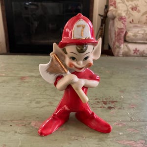 May include: A vintage ceramic figurine of a firefighter elf. The elf is wearing a red uniform and a red helmet with the number 7. The elf is holding a gold and white axe. The figurine has a smiling face and large ears.