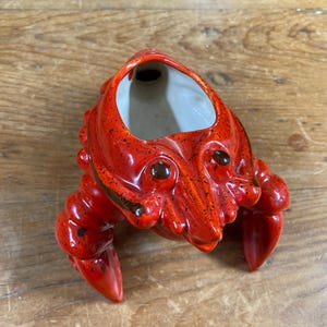 May include: A glossy ceramic crab-shaped vessel featuring a vibrant red glaze with dark speckles. The interior is finished in a smooth white glaze, and the exterior is molded with detailed claws, legs, and eyes, creating a functional container.