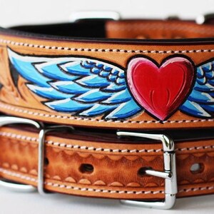 May include: A brown leather dog collar with a hand-painted design of blue wings and a red heart. The collar has a silver buckle and is stitched with white thread.