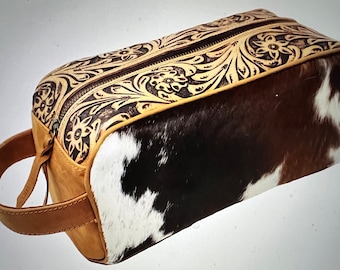 May include: A brown leather travel bag with a floral design and a cowhide patch. The bag has a zip closure and a handle.