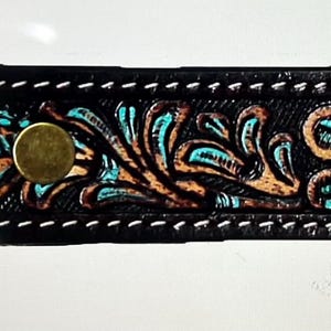 Puede incluir: Llavero de cuero negro con un diseño floral en turquesa y marrón. El llavero tiene un cierre de metal y un botón de latón. Los bordes están cosidos con hilo blanco.