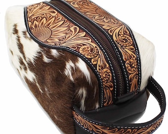 May include: A rectangular toiletry bag featuring brown and white cowhide panels on the sides and a central strip of tan leather with intricate floral tooling. The bag includes a dark brown zip closure and a matching leather carrying handle.