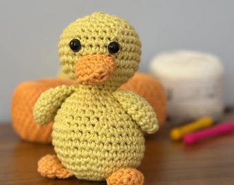 May include: A handmade crochet duckling in yellow and orange. The duckling has black safety eyes and an orange beak and feet. The duckling is sitting on a wooden surface. In the background are orange and white yarn balls.
