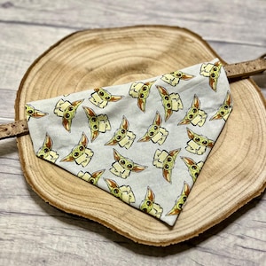 Könnte beinhalten: Ein dreieckiges Haustierhalstuch mit einem sich wiederholenden Muster aus kleinen grünen Alien-Figuren mit großen Ohren und dunklen Augen auf hellgrauem Grund, befestigt an einem braunen, strukturierten Halsband.