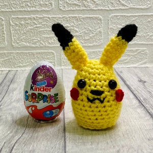May include: A yellow crocheted Pikachu toy with black ears, red cheeks, and black eyes. The toy is next to a Kinder Surprise egg.