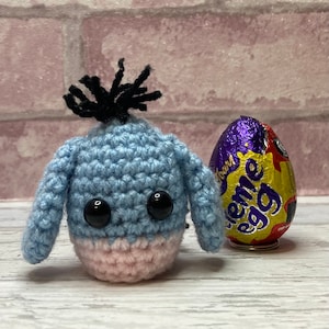May include: A blue crocheted Eeyore character with black yarn hair and pink ears. The character is sitting next to a purple and gold foil-wrapped chocolate egg.