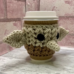 May include: A crocheted coffee cup cosy in the shape of a Yoda character from Star Wars. The cosy is beige and brown with black eyes.