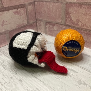 Könnte beinhalten: Eine gehäkelte Figur von Venom in Schwarz und Weiß mit einer roten Zunge, neben einer Terry's Chocolate Orange, die in goldene Folie eingewickelt ist. Die Schokoladenorange trägt den Text "Terry's Chocolate Orange" und "Made with Real Orange Oil".