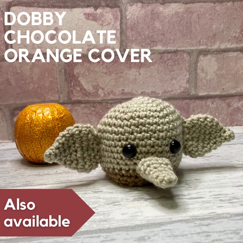 K&ouml;nnte beinhalten: Eine geh&auml;kelte Dobby-Haus-Elfenpuppe mit gro&szlig;en Ohren, braunen Augen und einer braunen Nase. Die Puppe sitzt auf einer wei&szlig;en Holzoberfl&auml;che neben einer verpackten orangen Schokolade. Der Text "Dobby Chocolate Orange Cover" befindet sich oben im Bild und der Text "Also available" unten im Bild.