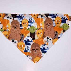 Könnte beinhalten: Ein weißes Hundehalstuch mit Star Wars-Motiv. Das Halstuch zeigt ein sich wiederholendes Muster von Figuren aus den Star Wars-Filmen, darunter Chewbacca, R2-D2, C-3PO, Yoda und andere. Das Halstuch ist an einem braunen Lederband befestigt.