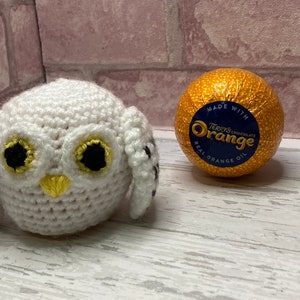 Könnte beinhalten: Weißer gehäkelter Eule mit schwarzen und gelben Details, neben einer goldenen Schokoladenkugel mit dem Text "Terry's Chocolate Orange" und "Made with Real Orange Oil".