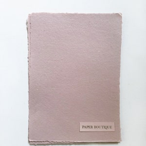 以下が含まれることがあります： 右下隅に「PAPER BOUTIQUE」と印刷されたラベルが付いた、薄いピンク色のテクスチャ加工された紙のシートです。