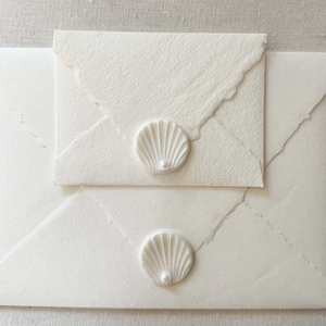 Op de afbeelding: Twee off-white enveloppen van gestructureerd papier met geschepte randen. Elke envelop is verzegeld met een ronde witte waszegel met een reliëf van een schelp en een kleine, centraal geplaatste witte parel op de envelopflap.