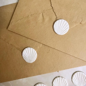 May include: Image of stationery elements: two brown paper envelopes and several white wax seals. The seals are round with a textured, fan-like design. The envelopes are slightly textured and have a natural, earthy tone.