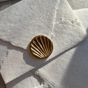 Op de afbeelding: Close-up van verschillende witte papieren enveloppen met een getextureerd oppervlak. Elke envelop is verzegeld met een goudkleurige waszegel. De zegels hebben een gedetailleerd, waaiervormig ontwerp. De enveloppen zijn gestapeld, met schaduwen die diepte toevoegen.