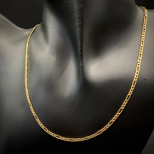 May include: A gold-colored Figaro chain necklace displayed on a black mannequin. The necklace features a pattern of two or three short links followed by a longer oval link. The chain is a bright, shiny gold color.