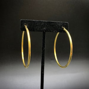 Pode incluir: Um par de brincos de argola dourados expostos em um suporte preto. Os brincos s&atilde;o simples e de forma circular.