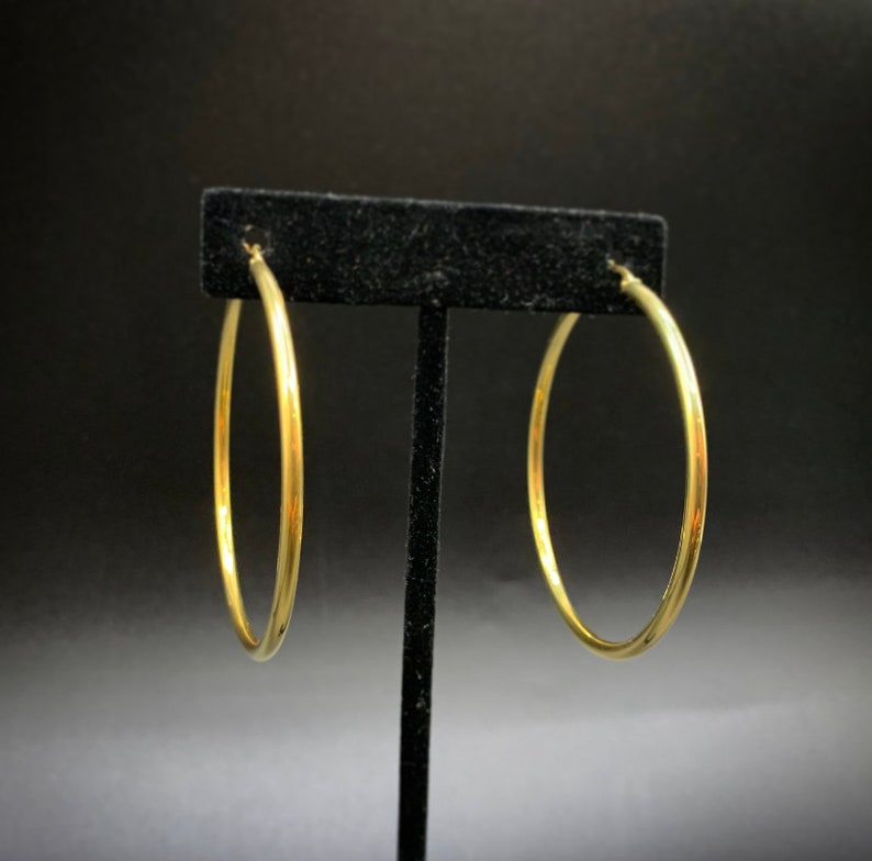 Pode incluir: Um par de brincos de argola dourados expostos em um suporte preto. Os brincos s&atilde;o simples e de forma circular.