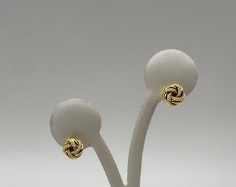 May include: A pair of gold-colored stud earrings. Each earring features a textured knot design. The earrings are displayed on white earring stands against a plain background.