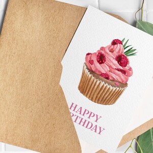 May include: A watercolor birthday card featuring a pink cupcake with raspberries and hearts. The card is inside a brown envelope. The words "HAPPY BIRTHDAY" are printed in pink on the card.