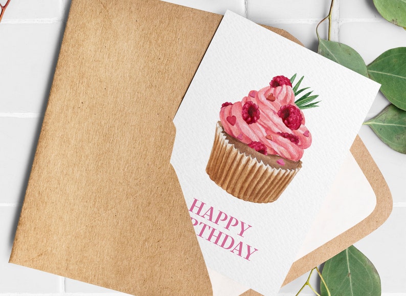 May include: A watercolor birthday card featuring a pink cupcake with raspberries and hearts. The card is inside a brown envelope. The words "HAPPY BIRTHDAY" are printed in pink on the card.