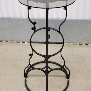 May include: A black metal side table with a round glass top. The table has a unique design with a gear-shaped pattern on the glass top and a circular base.
