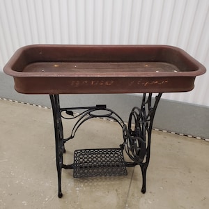 May include: A vintage metal planter with a rusty brown finish and black metal legs. The planter is shaped like a rectangular trough and has a decorative design on the legs. The planter is sitting on a light gray floor.