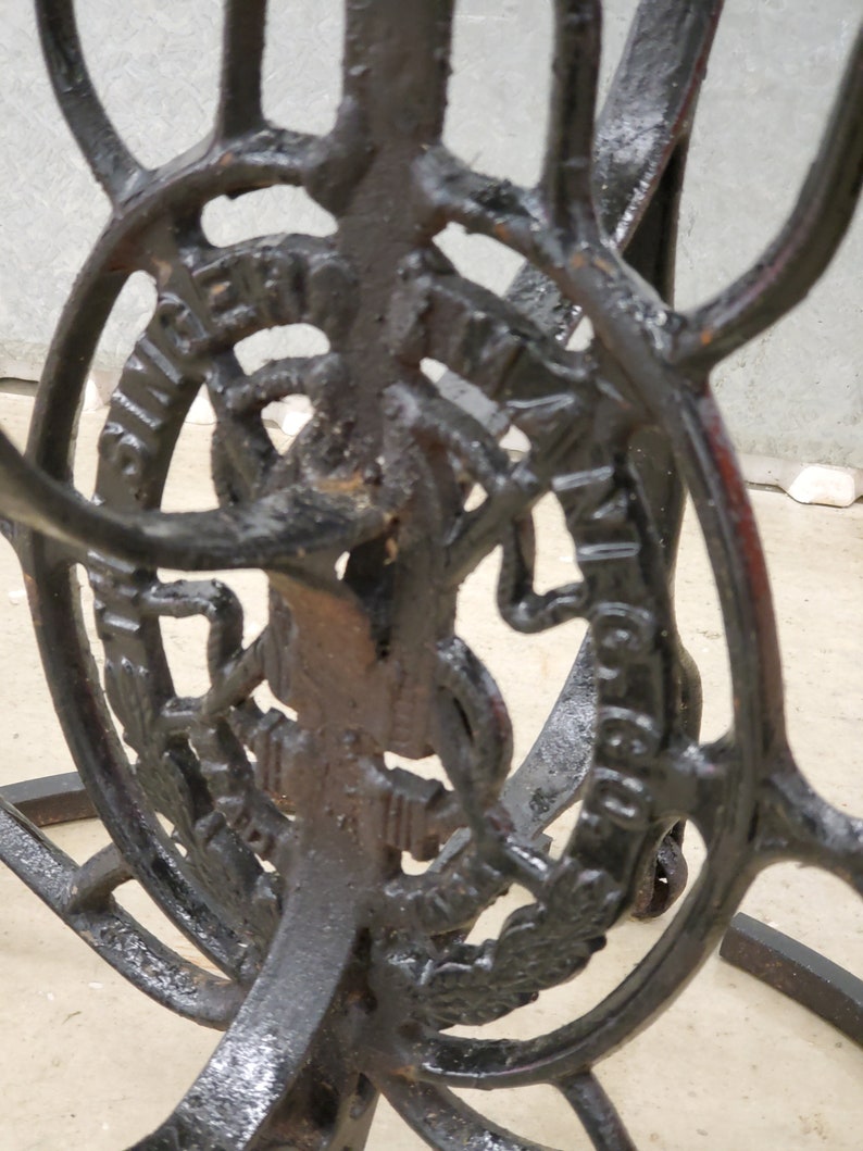 May include: Black metal stand with intricate details and a circular design featuring the text "Singer Manufacturing Co."