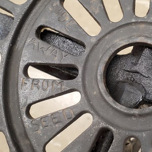 May include: Close-up of a black metal wheel with the words "AWAY FROM SEED" inscribed on it. The wheel has a series of evenly spaced holes around its circumference.