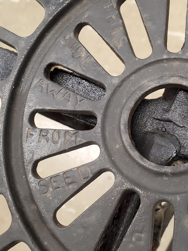 May include: Close-up of a black metal wheel with the words "AWAY FROM SEED" inscribed on it. The wheel has a series of evenly spaced holes around its circumference.