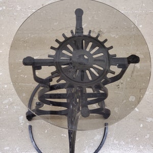May include: Black metal side table with a round glass top and a decorative gear design. The table has a unique, spiral base.