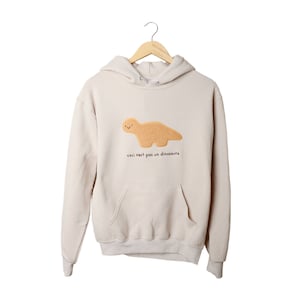 May include: A beige hoodie with a brown dinosaur-shaped cookie on the front. The text "Ceci n'est pas un dinosaure" is printed below the cookie.