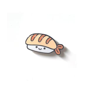 May include: A small, cartoon-style enamel pin of a sushi roll with a smiling face. The sushi is orange and white with a red and white seaweed wrap.