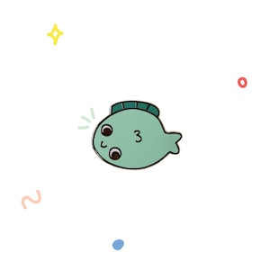 May include: A green enamel pin shaped like a fish with a number "3" on its side. The fish has large eyes and a smiling mouth.