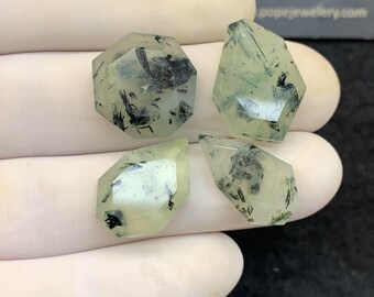Könnte beinhalten: Vier facettierte Prehnit-Edelsteine mit durchscheinenden hellgrünen Farbtönen und dunklen Einschlüssen. Die Edelsteine werden in einer behandschuhten Hand gehalten, mit dem Text "POPE JEWELRY" im Hintergrund.