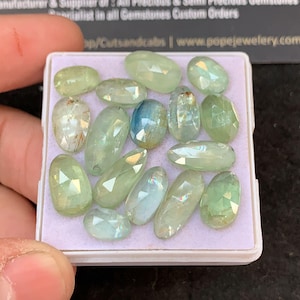 May include: A collection of faceted gemstones in various shapes and sizes, primarily in shades of light green and a few with blue hues. The gems are displayed in a white rectangular tray, showcasing their clarity and cut. The text "POPE JEWELRY" is visible in the background.