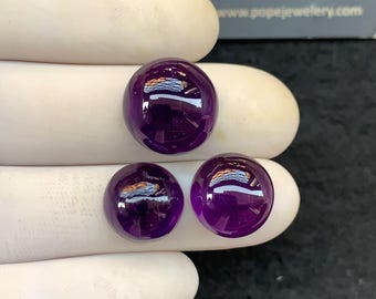 May include: Three polished, round, deep purple gemstones. The gems are smooth and reflective, with a high lustre. The image is taken from a close-up perspective, highlighting the colour and clarity of the stones. The background is a neutral colour.